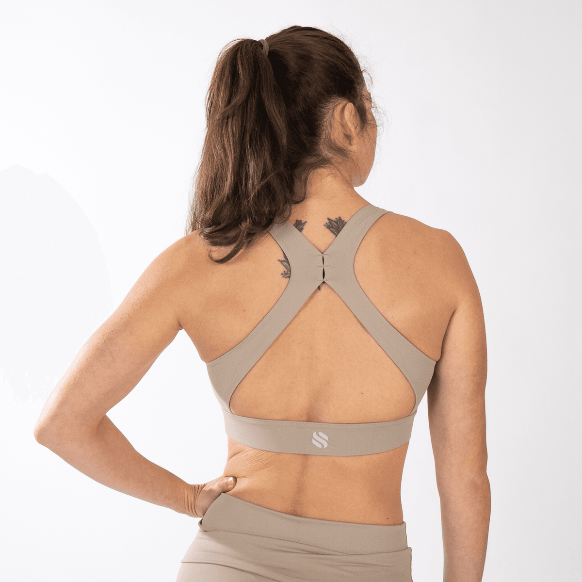 A beige sports bra with a racerback strap design and a small logo on the back band worn by a person from behind.