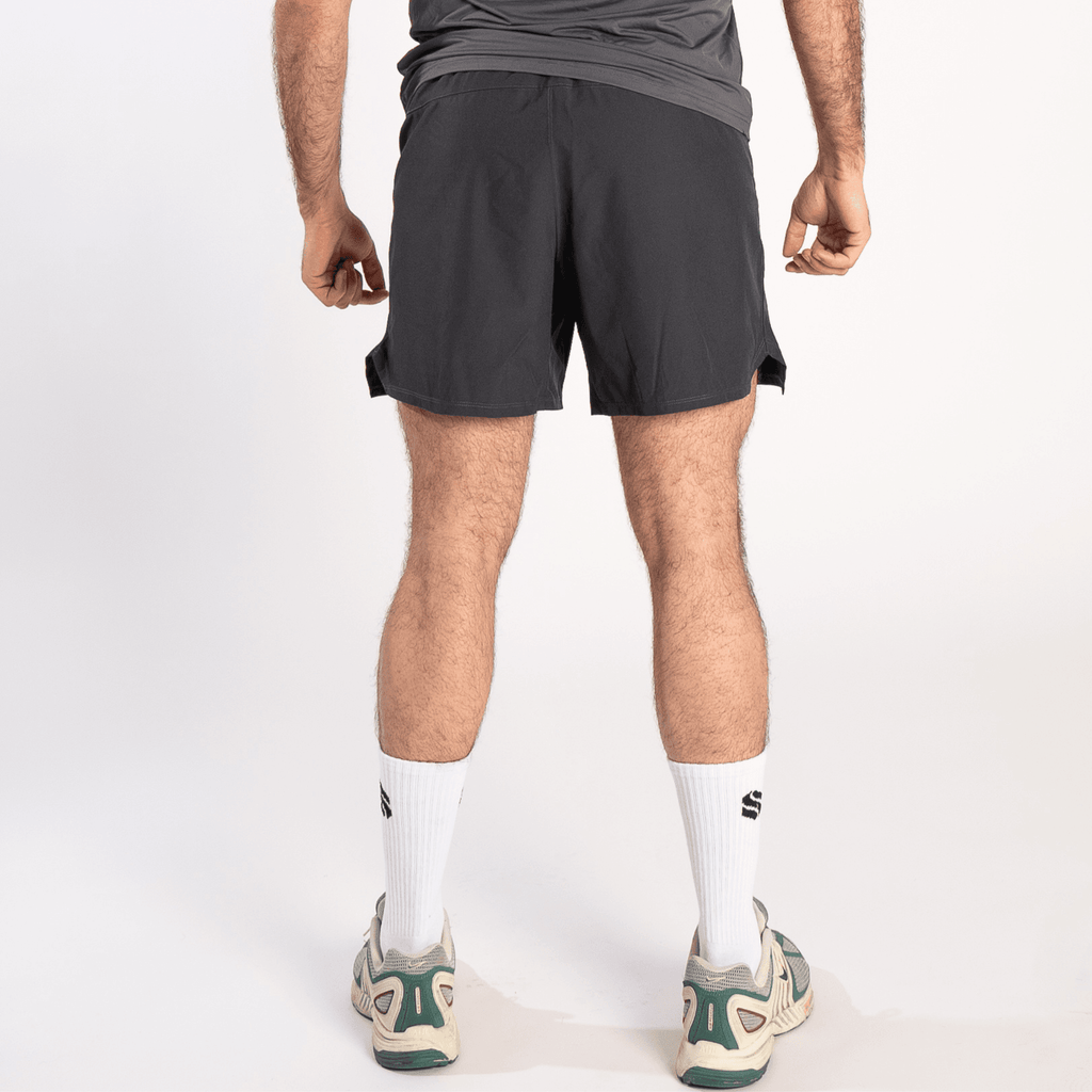 A person viewed from the back wears grey athletic shorts, white crew socks with a small logo, and grey and green running sneakers against a plain white background.