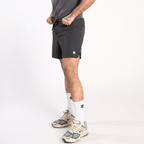 A person wearing dark grey athletic shorts with a small white circular logo on the left thigh and light-colored sneakers with green accents.