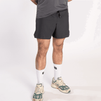 A person wears charcoal grey athletic shorts with a drawstring waist, white crew socks with a black logo, and cream, green, and yellow sneakers against a plain white background.