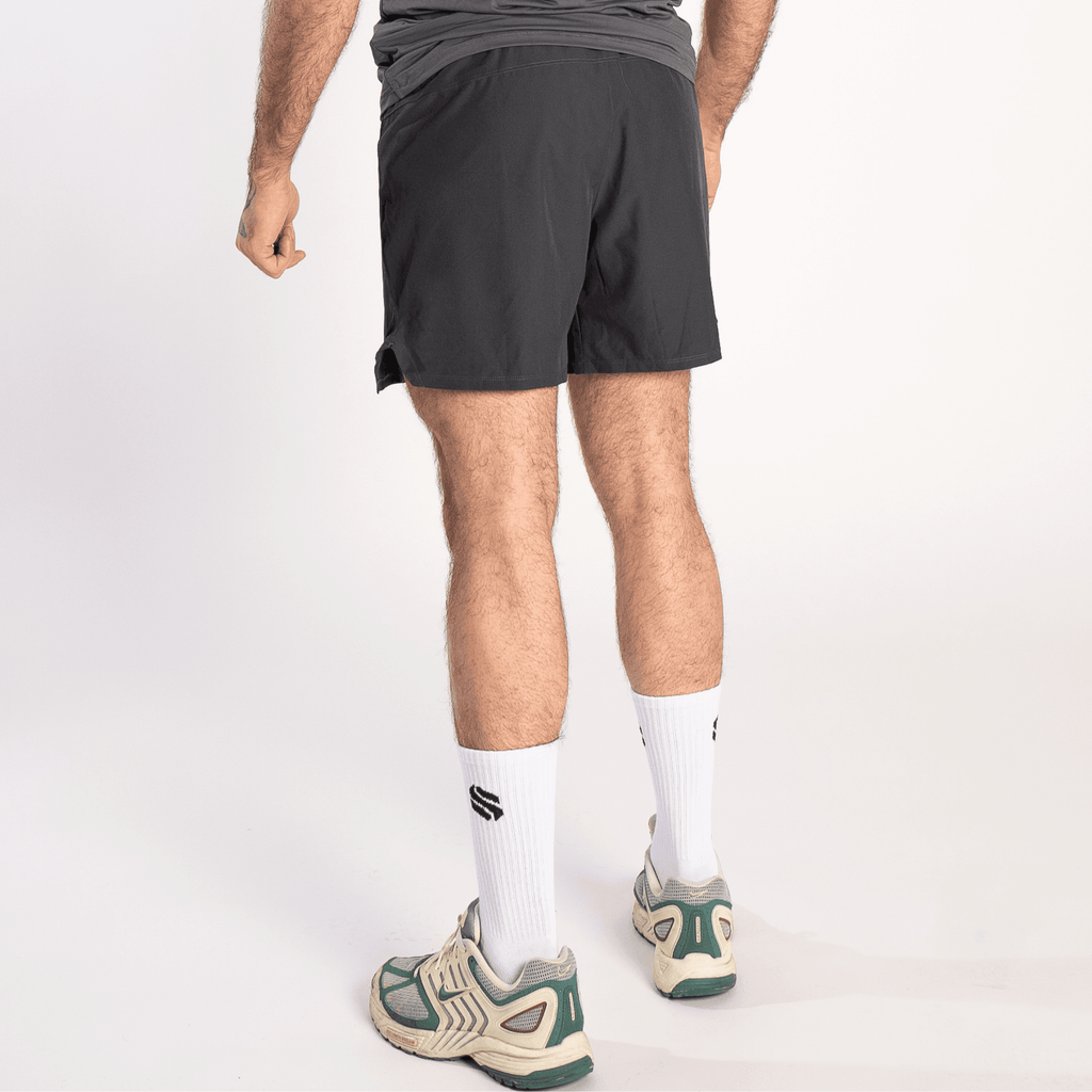 A person viewed from behind wearing black athletic shorts and white crew socks with a black logo, paired with beige, green, and white sneakers.