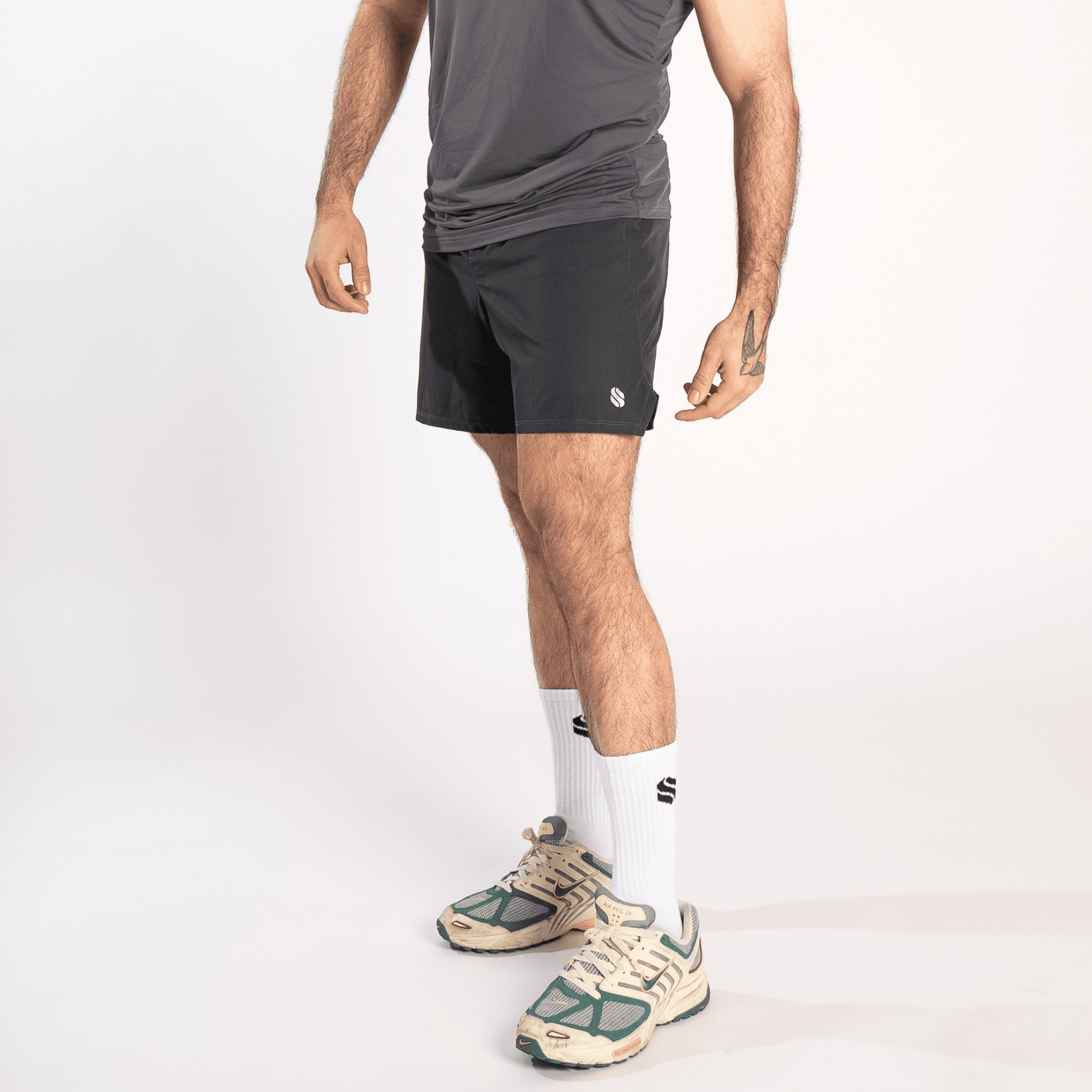 A man wearing a grey athletic tank top, dark grey shorts, white crew socks, and beige and green athletic sneakers against a white background.