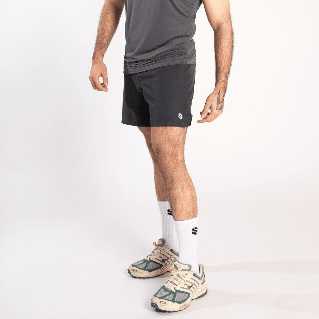 A man wearing a grey athletic tank top, dark grey shorts, white crew socks, and beige and green athletic sneakers against a white background.