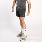 A man wearing a grey athletic tank top, dark grey shorts, white crew socks, and beige and green athletic sneakers against a white background.