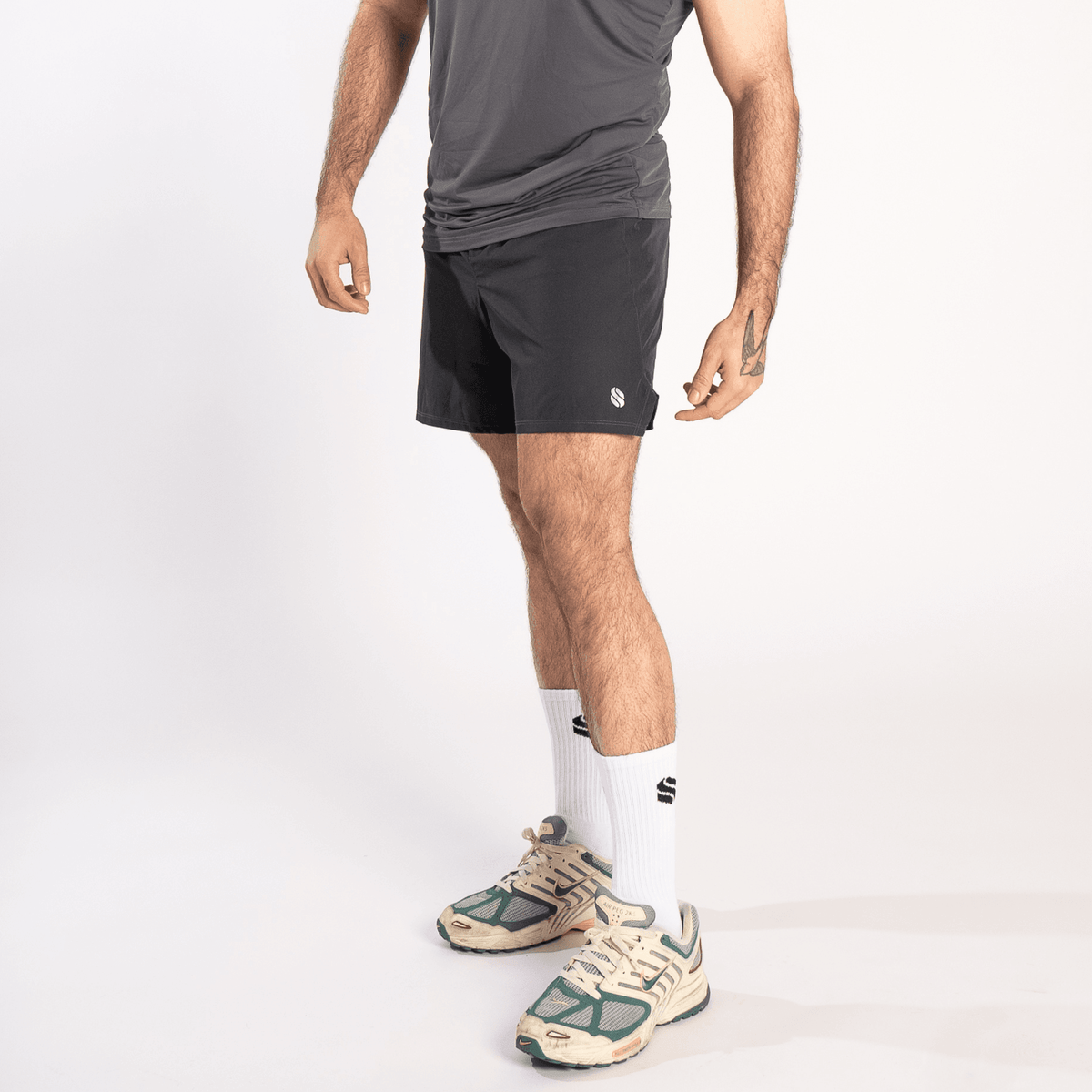 A man wearing a grey athletic tank top, dark grey shorts, white crew socks, and beige and green athletic sneakers against a white background.