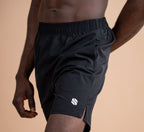 A close-up of a man wearing black athletic shorts with a white logo on the lower left leg, set against a solid warm beige background.