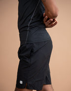 A close-up side view of a person wearing black athletic shorts and a matching black top against a solid warm beige studio background. A small white logo is visible on the lower left hem of the shorts.