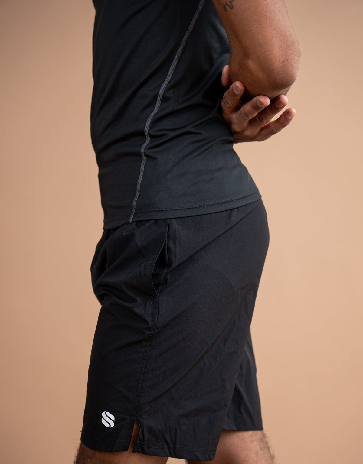 A close-up side view of a person wearing black athletic shorts and a matching black top against a solid warm beige studio background. A small white logo is visible on the lower left hem of the shorts.