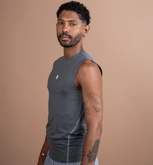 A man wearing a dark gray sleeveless athletic tank top and light gray shorts, standing against a solid warm tan background.