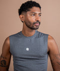 A man with short curly black hair and a beard wearing a gray sleeveless athletic shirt with a small white logo on the chest, photographed against a solid warm beige background.