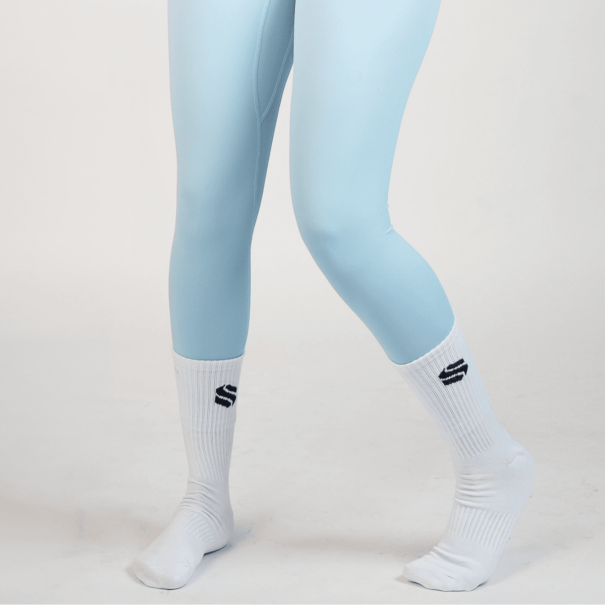 A pair of light blue leggings worn with white mid-calf ribbed socks featuring a black geometric logo on a plain white background.