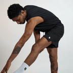 A man wearing a black sleeveless athletic shirt and black athletic shorts stretches his leg forward against a plain white background.