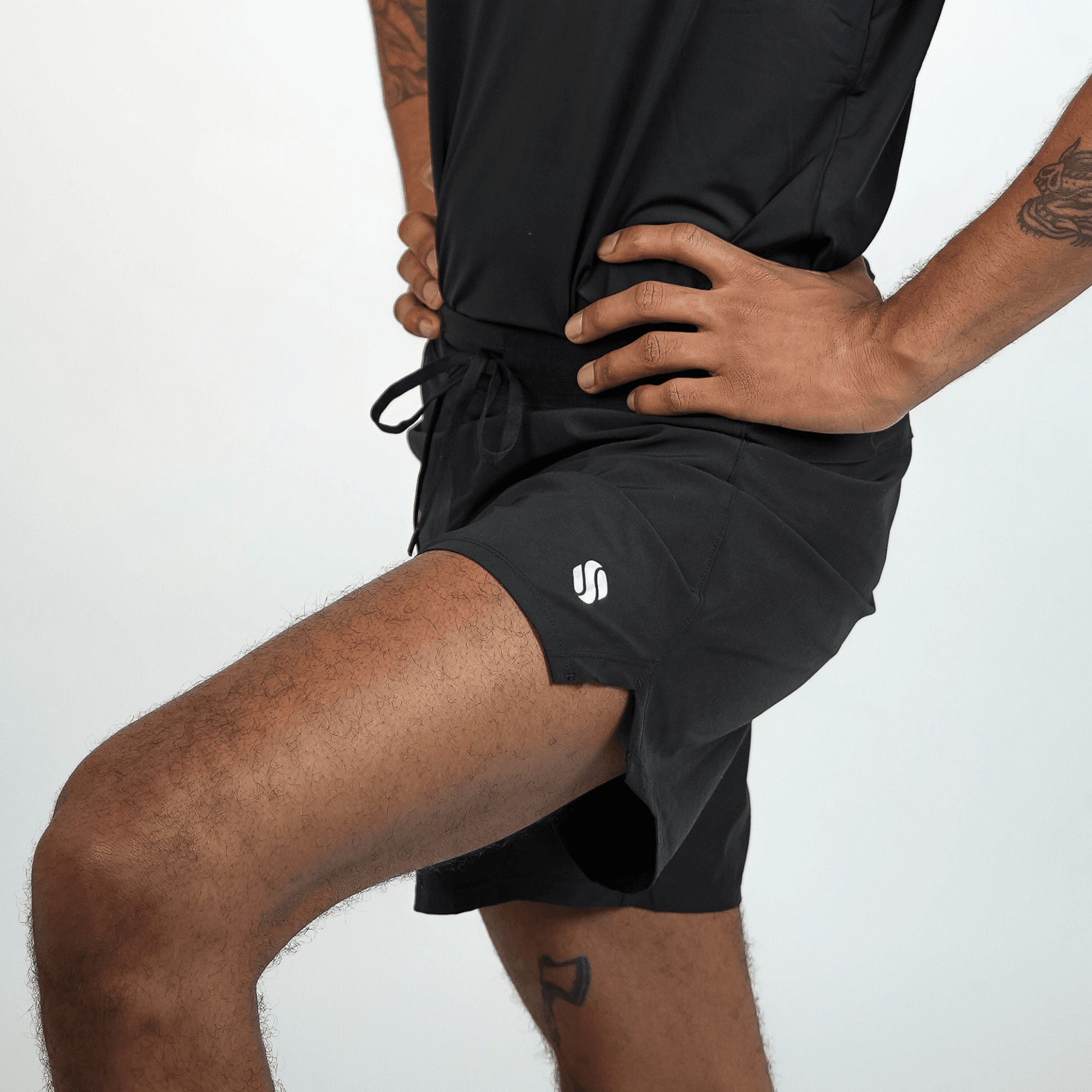 Black athletic shorts with an elastic drawstring waist and a small white circular logo on the right side are worn by a man with a raised leg.