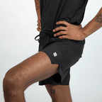 Black athletic shorts with an elastic drawstring waist and a small white circular logo on the right side are worn by a man with a raised leg.