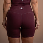 Rear view of a woman wearing a matching burgundy activewear set with high-waisted shorts and a strappy top, featuring a small white stylized 'S' logo on the back of both pieces, set against a solid warm brown background.