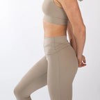 A side view of a person wearing high-waisted, beige leggings and a matching sports bra against a white background.