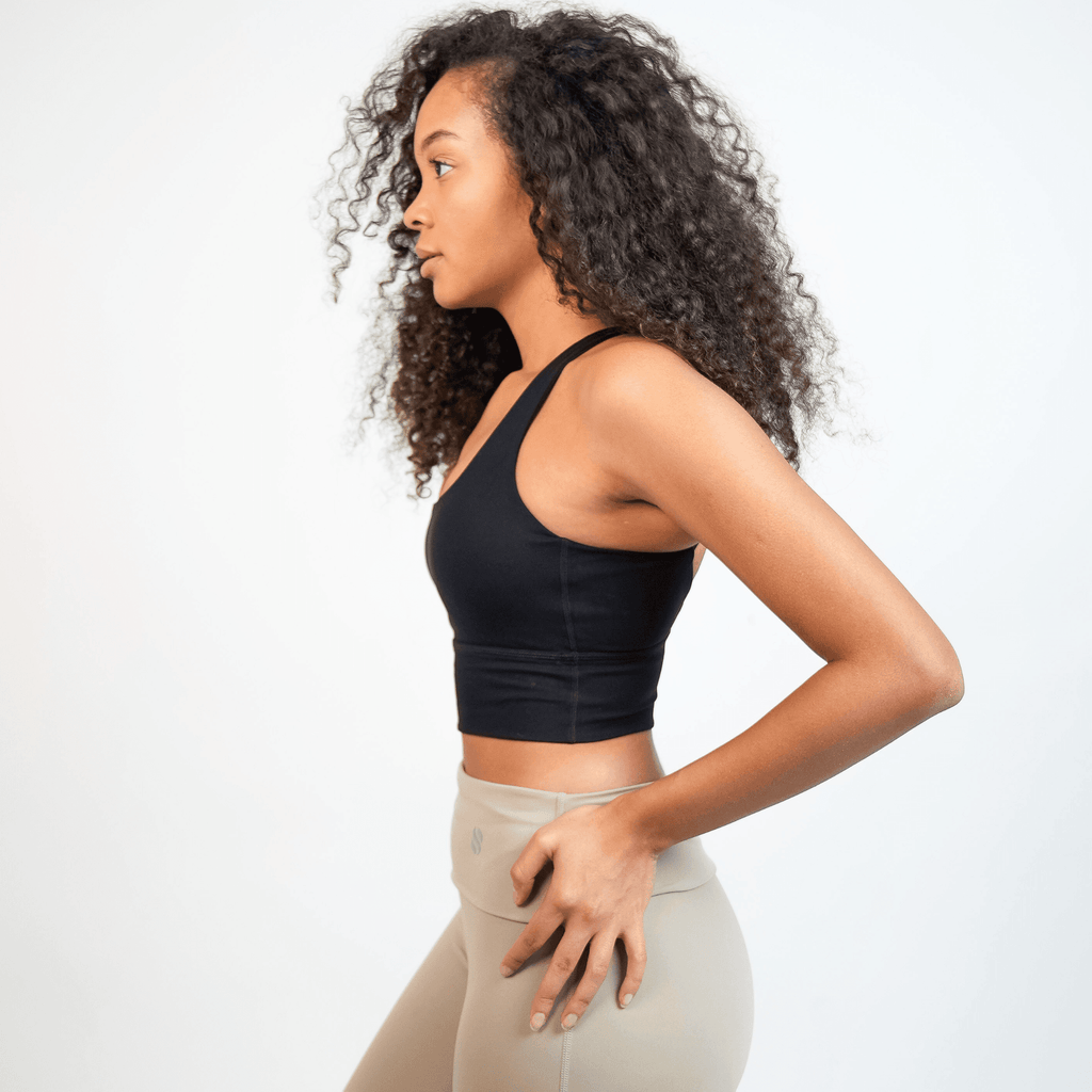 A woman with curly hair wears a black cropped sports bra paired with high-waisted beige leggings against a plain white background.