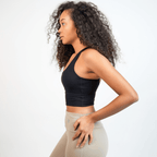 A woman with curly hair wears a black cropped sports bra paired with high-waisted beige leggings against a plain white background.